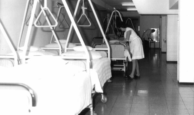Historisches Bild eines Krankenhausflures, in dem einige Patientenbetten stehen und eine Krankenpflegerin eins der Betten richtet. Im Hintergrund ist ein Arzt, welcher sich bückt.