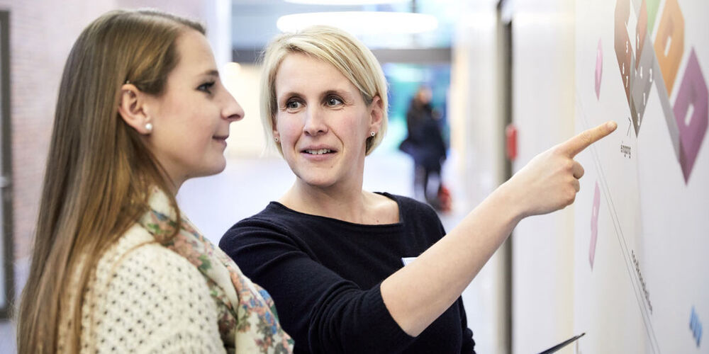 Zwei Frauen stehen vor einem Plakat und unterhalten sich über dieses. Die eine Frau zeigt der anderen auf diesem etwas mit ihrem Finger.