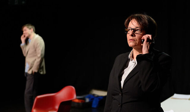 Theaterbühne auf der eine Frau im Vordergrund ist und ein Telefon ans Ohr hält. Im Hintergrund steht ein Mann und hat ebenfalls ein Telefon am Ohr. Zwischen ihnen befinden sich zwei Stühle.